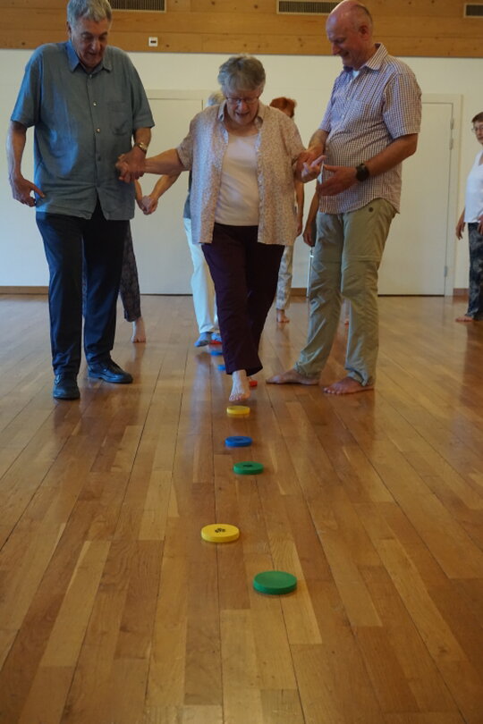 Seniorin hält sich an einem Stuhl fest und macht eine einfache Balance-Übung.